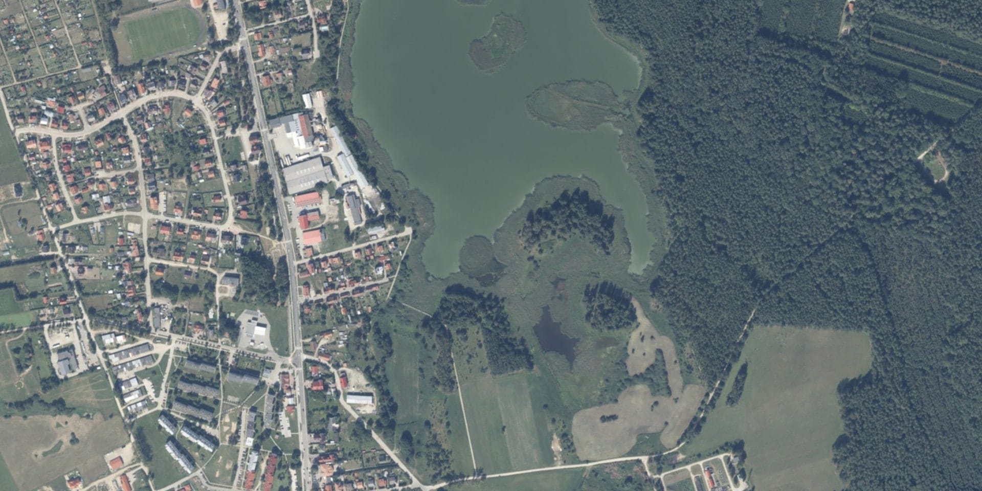 Staw w Pieckach z wyspami między zabudową wsi a lasem, widok lotniczy