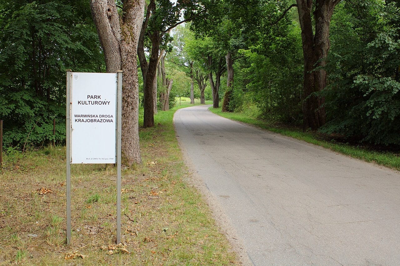 Asfaltowa droga wśród drzew i tablica Warmińskiej Drogi Krajobrazowej w Worytach